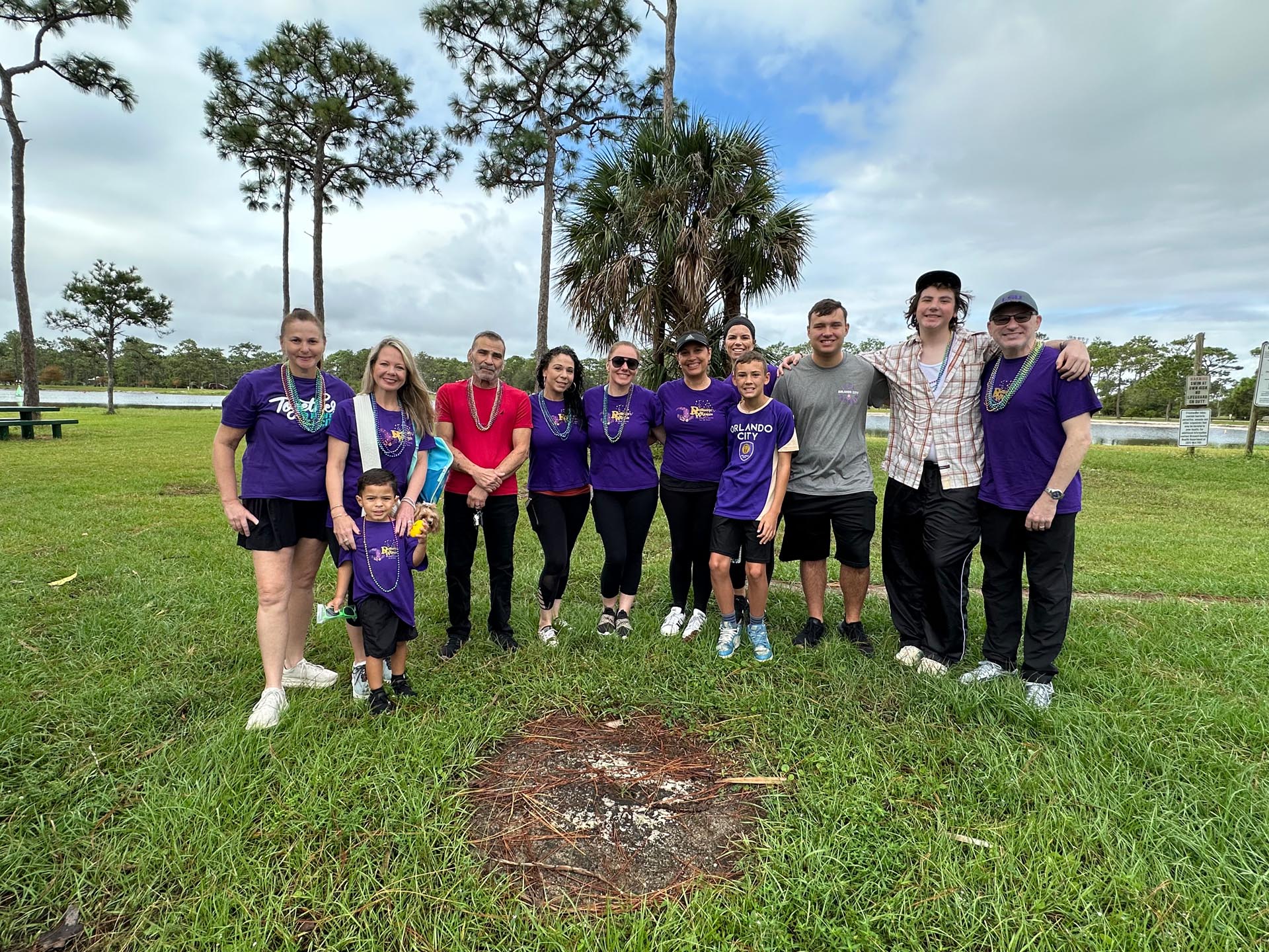 A group of eleven people and one child stand together on grass in a park, some wearing purple shirts, with trees and a body of water in the background.