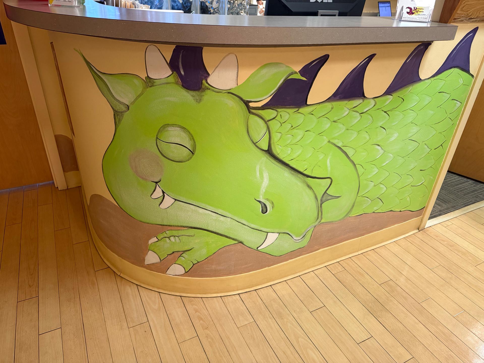 A painted mural of a green sleeping dragon with purple spikes decorates the front of a reception desk with a wood floor.