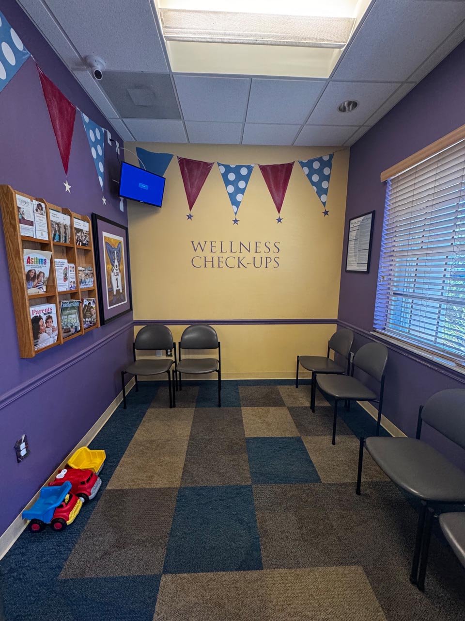 A small waiting room with empty chairs, children’s toys on the floor, bulletin boards with notices, and a wall sign reading “WELLNESS CHECK-UPS.”.