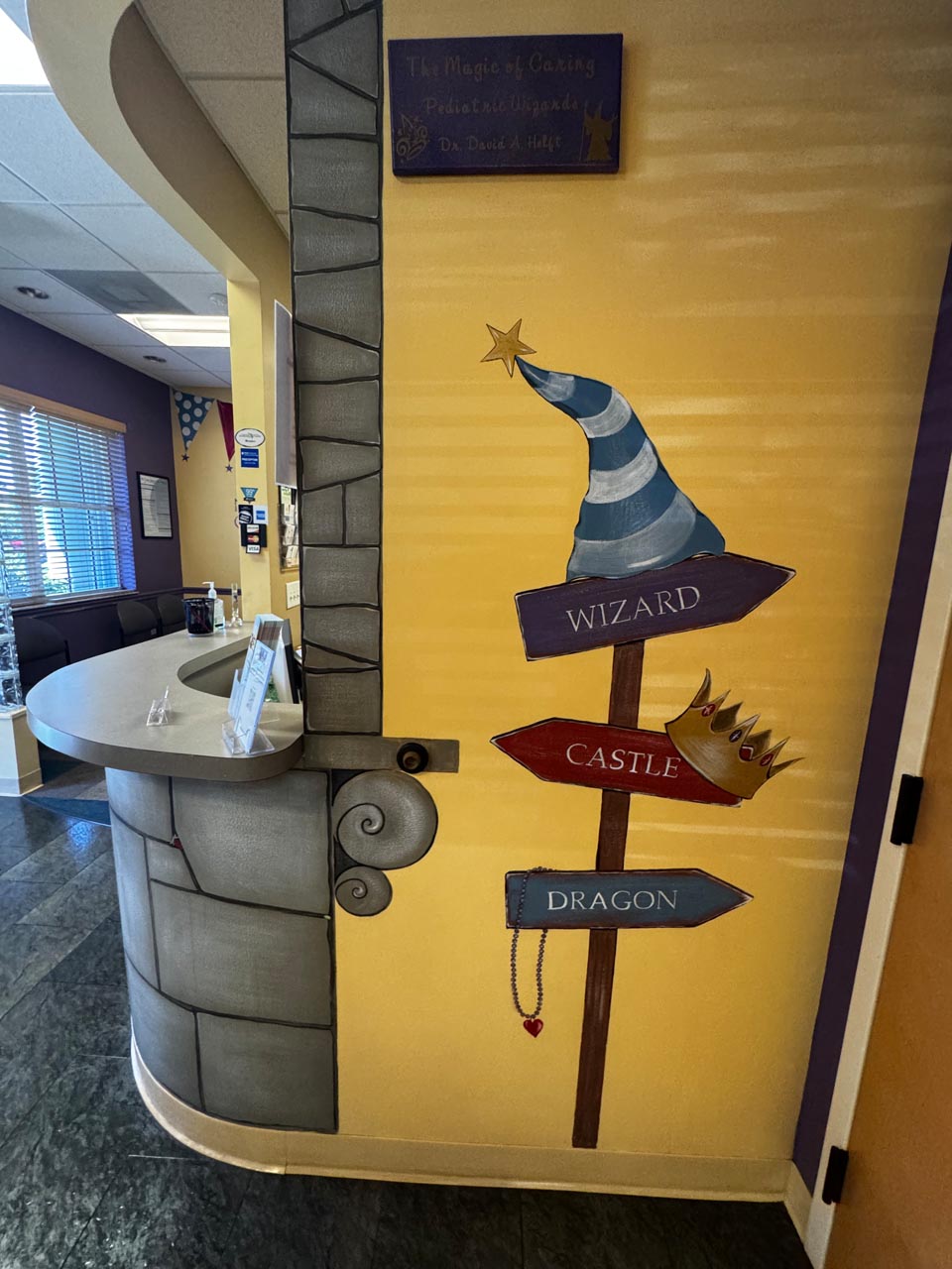 A whimsical wall mural in a pediatric office features a wizard hat, a crown, and directional signs labeled "Wizard," "Castle," and "Dragon" near a curved reception desk.
