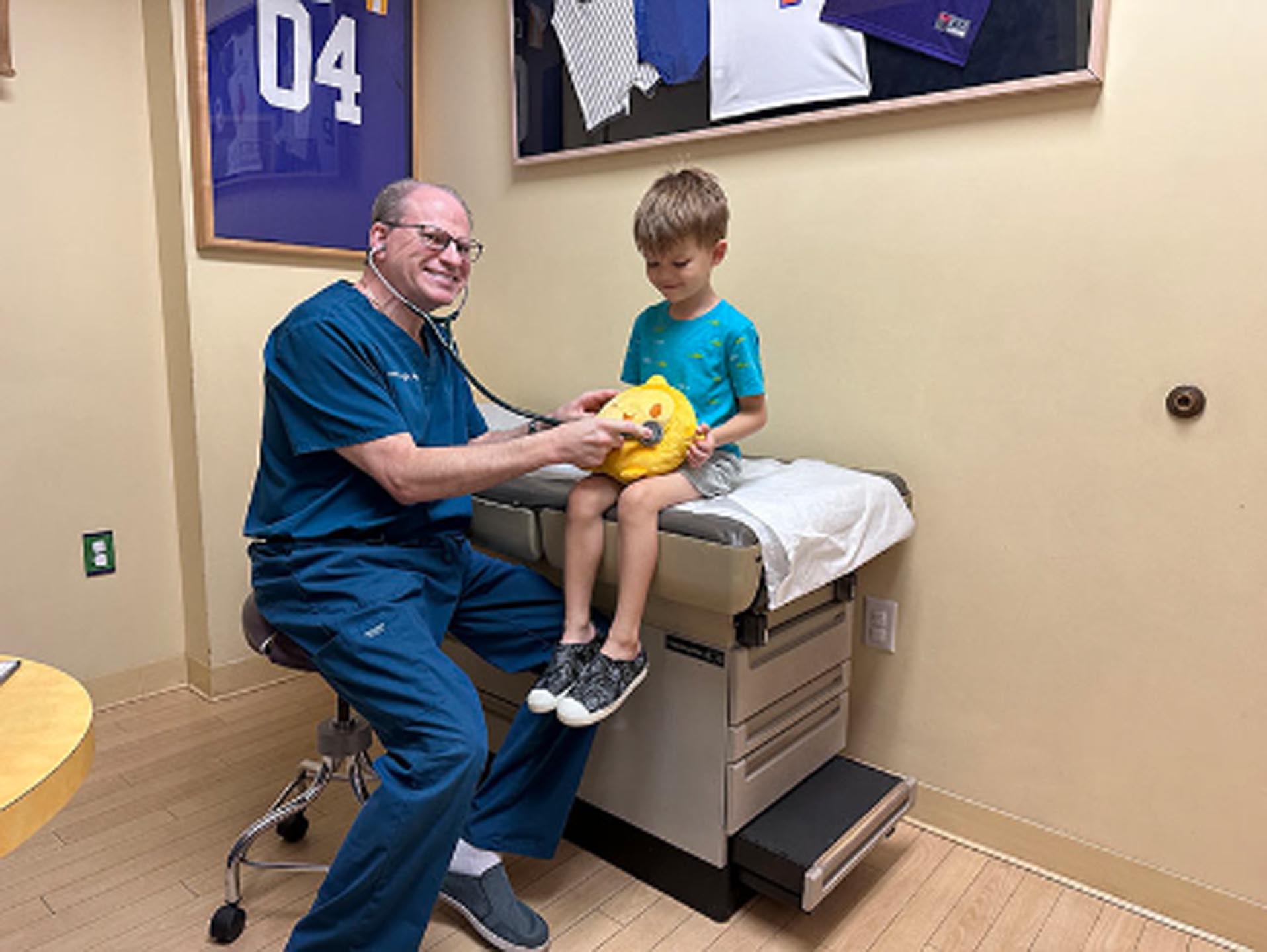 Pediatric Wizards: A Pediatrician in Melbourne, FL, David A. Helft, M.D., wearing blue scrubs uses a stethoscope on a plush toy held by a young boy sitting on an exam table in a medical office.