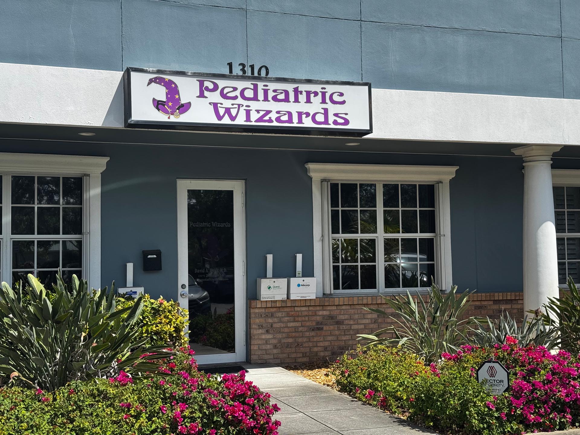 Front entrance of a building with a sign reading "Pediatric Wizards," surrounded by flowering plants and shrubs.