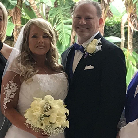 A bride in a white wedding dress holds a bouquet of white roses and stands beside a groom in a suit with a bow tie, both smiling in front of greenery.