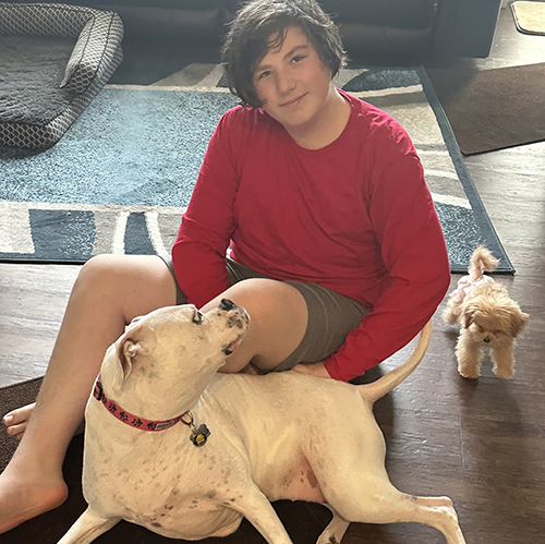 A boy in a red shirt sits on the floor with a large white dog lying beside him and a small tan puppy standing nearby.