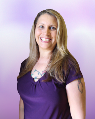 Smiling woman with long blonde hair wearing a purple shirt, showing tattoos on her chest and upper left arm, standing against a light purple background.