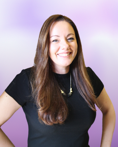 Woman with long brown hair wearing a black top and necklace stands smiling with hands on hips against a purple gradient background.