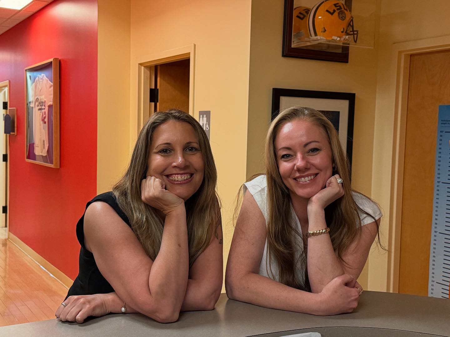 Two women with long hair are smiling and leaning on a counter in an office hallway decorated with sports memorabilia, creating a warm environment focused on adolescent health.