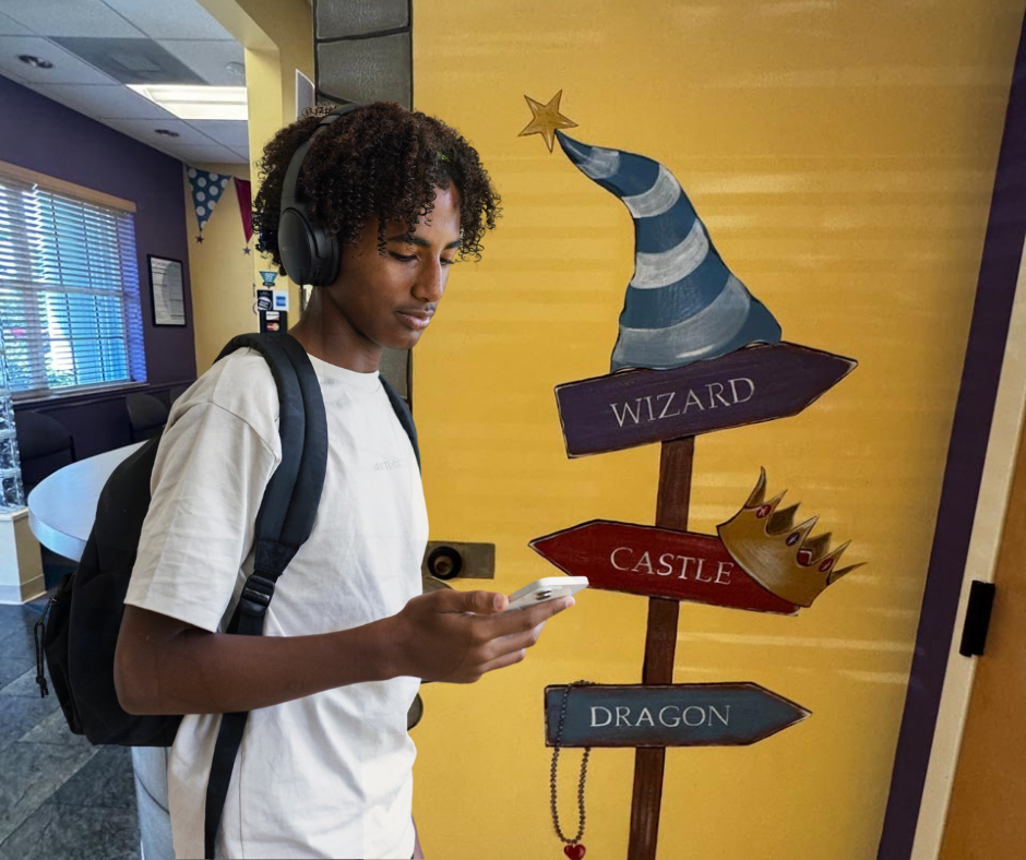 A young person with headphones and a backpack, focused on their phone, stands near a yellow wall adorned with whimsical signs reading "Wizard," "Castle," and "Dragon"—a snapshot that captures the spirit of teen health and curiosity.