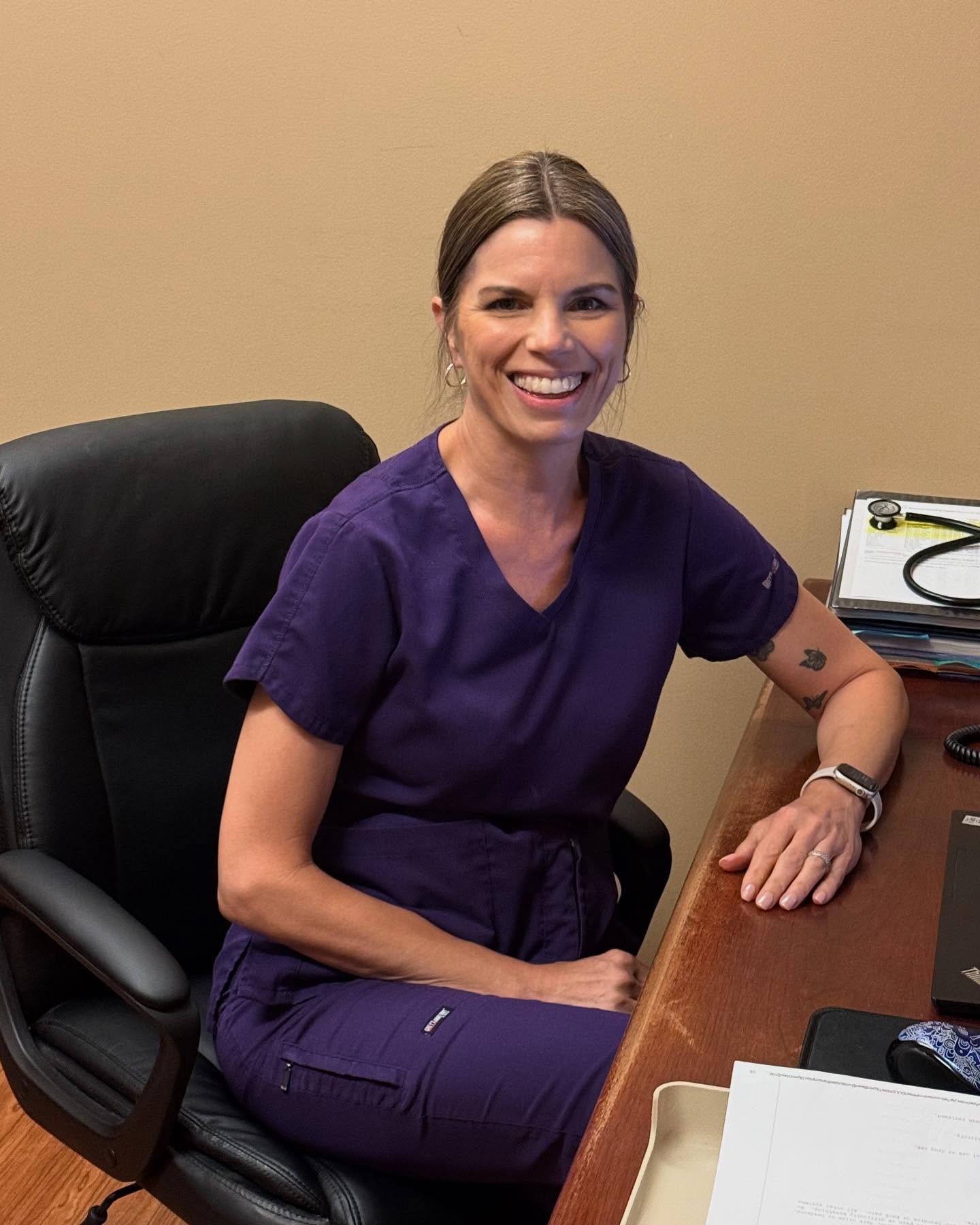 A woman in purple scrubs sits at a desk, smiling at the camera. Office items, paperwork, and a stethoscope are visible on the desk, reflecting her dedication to adolescent health.