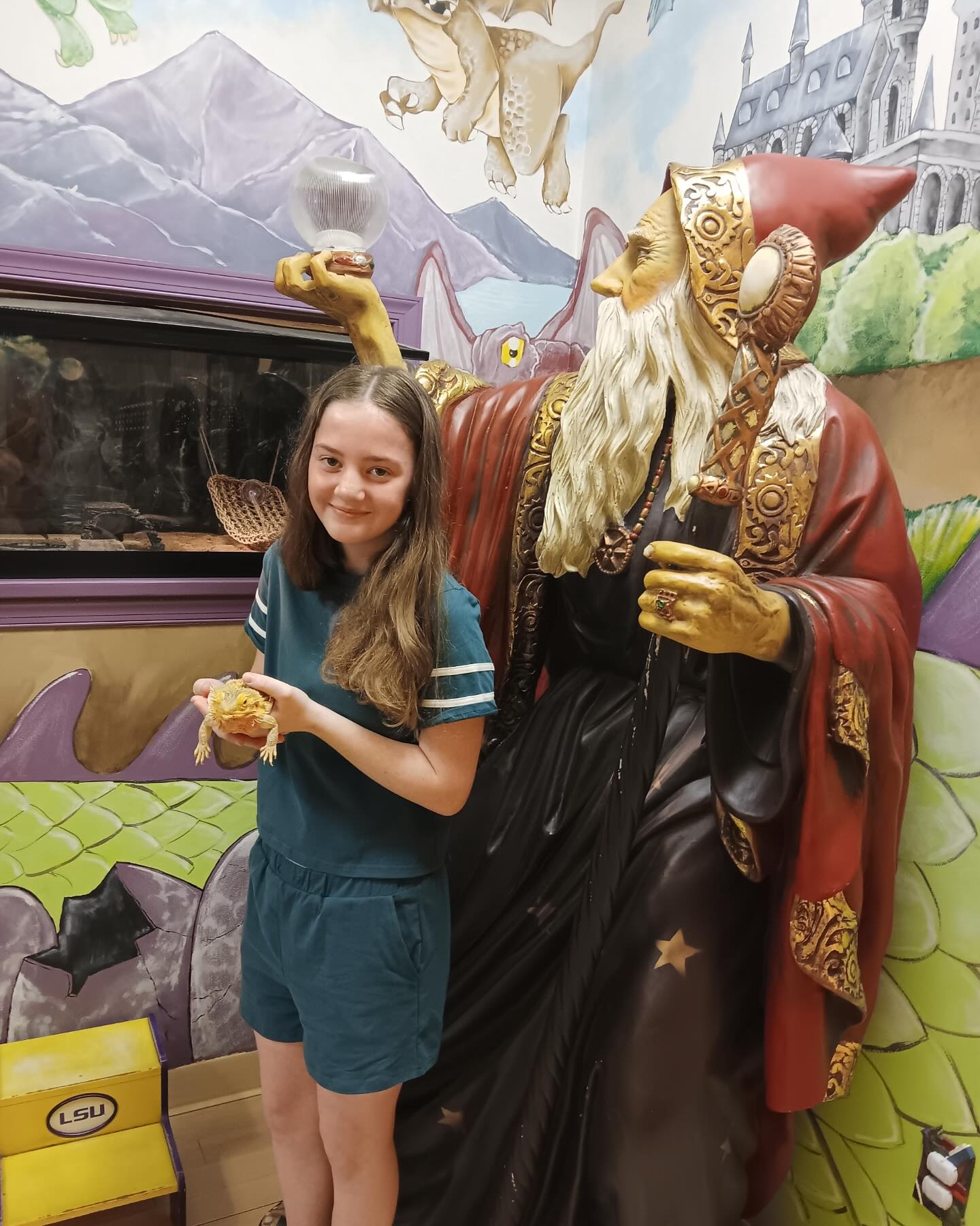 Adolescent Health Services in Melbourne, FL: Pediatric Wizards. A girl stands next to a large wizard statue, holding a lizard; the background features a painted castle and dragons, capturing a magical moment that celebrates youth health and imagination.