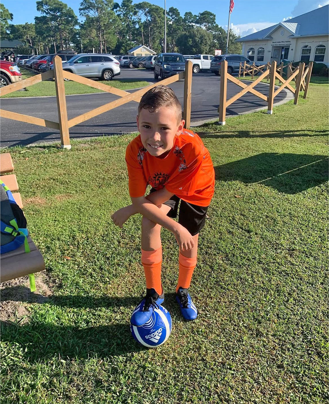 A boy in an orange soccer uniform stands on grass with one foot on a blue soccer ball, smiling at the camera, ready for his sports school physicals.