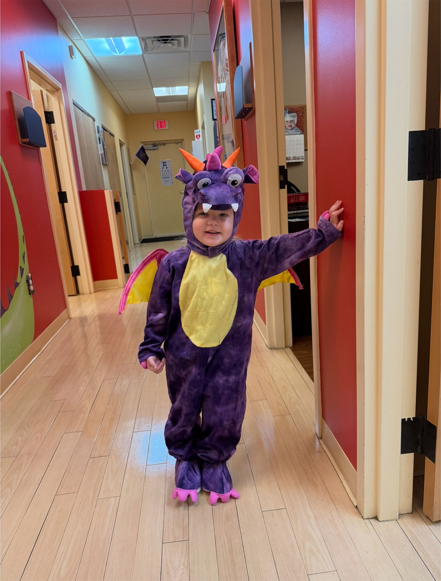 A young child stands in a hallway, smiling at the camera in a purple dragon costume with yellow wings and belly—ready for fun after their vaccination in Melbourne, FL.