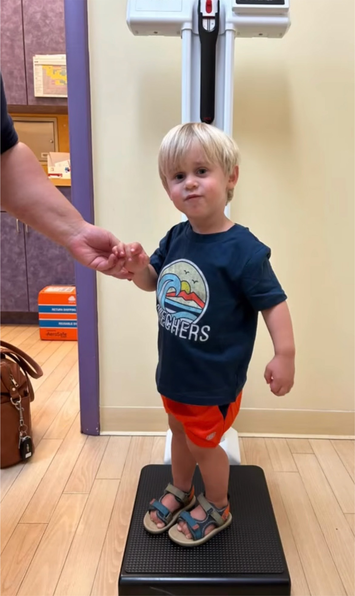 A young child stands on a medical scale, holding an adult's hand, in a clinic setting—possibly waiting for vaccination in Melbourne, FL—with a purse and colorful boxes in the background.