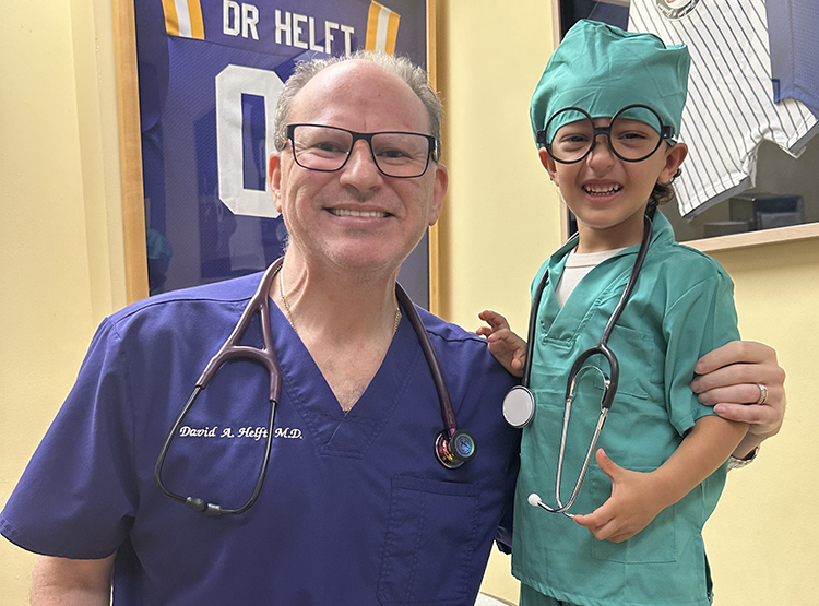 An adult in medical scrubs stands smiling with a young child dressed in a green doctor costume, both wearing stethoscopes. Sports jerseys are displayed in the background.