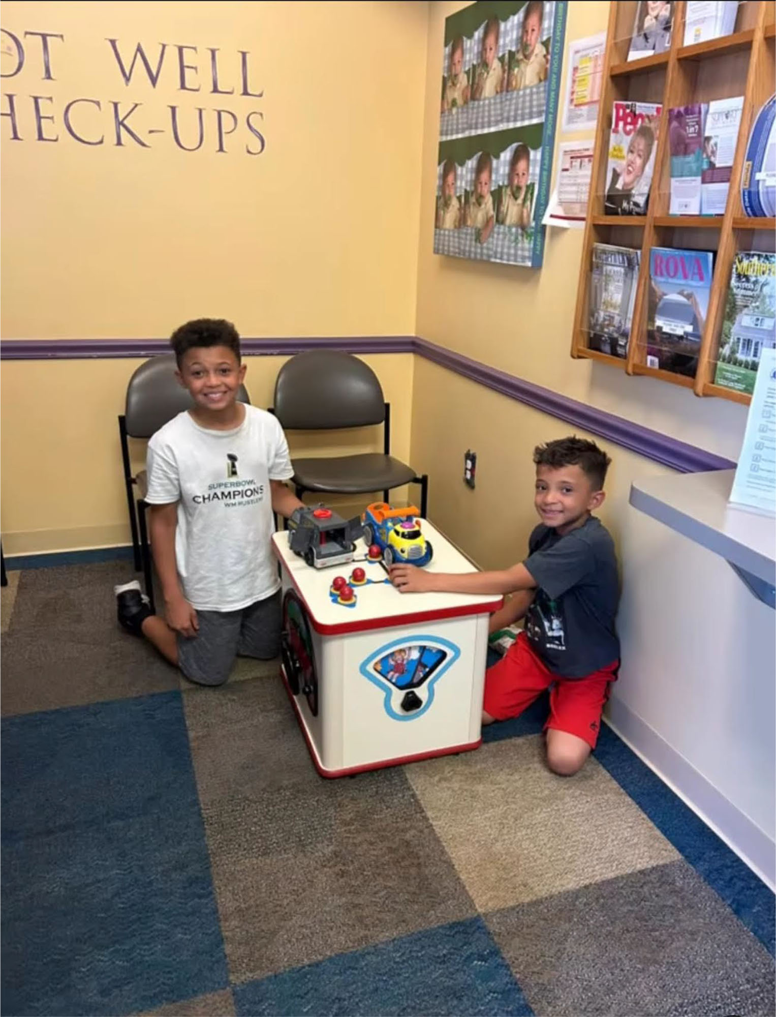 Two boys are kneeling and playing with a toy activity table in the waiting room of a pediatrician in Melbourne, FL. Chairs, a magazine rack, and a “Well Check-Ups” wall sign are visible in the background.