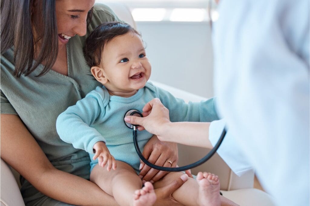 A smiling baby sits on a woman's lap while a healthcare professional listens to the baby's chest with a stethoscope, as the mother reviews insurance information.