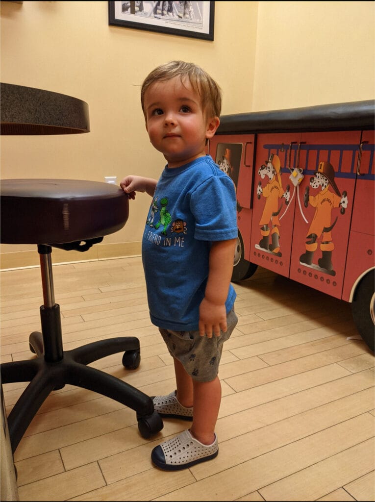 A young child stands in a room with wood flooring, next to a stool and a cabinet decorated with firefighter illustrations, ready for well-child checkups.