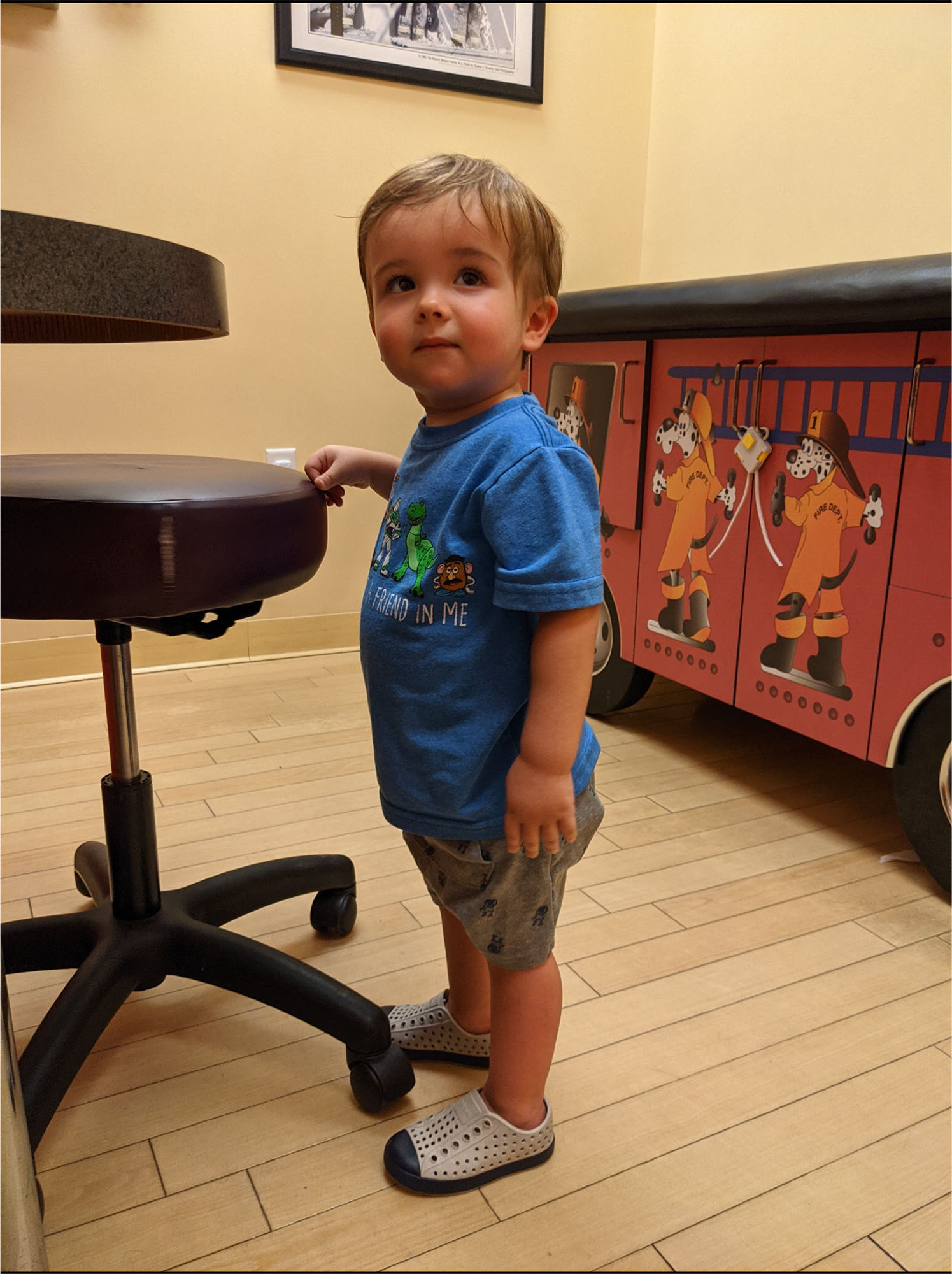 A young child stands in a room with wood flooring, next to a stool and a cabinet decorated with firefighter illustrations, ready for well-child checkups.