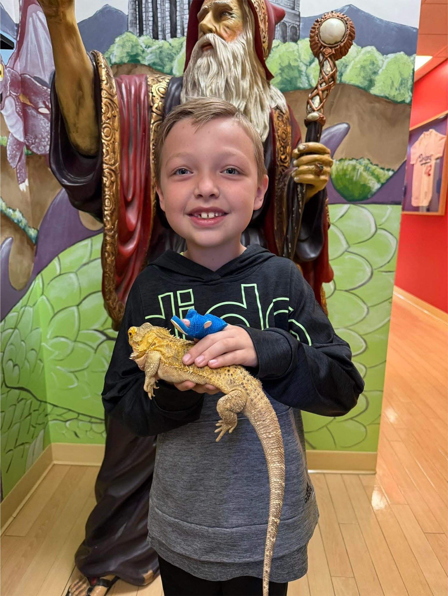 A smiling child holds a bearded dragon and a small blue toy, standing in front of a wizard statue in the brightly colored office of a pediatrician in Melbourne, FL.