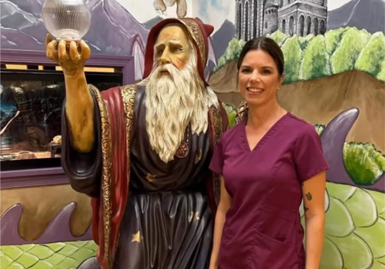 A woman in purple scrubs stands next to a statue of a wizard holding a crystal ball, with a colorful mural in the background—highlighting adolescent health in Melbourne, FL.
