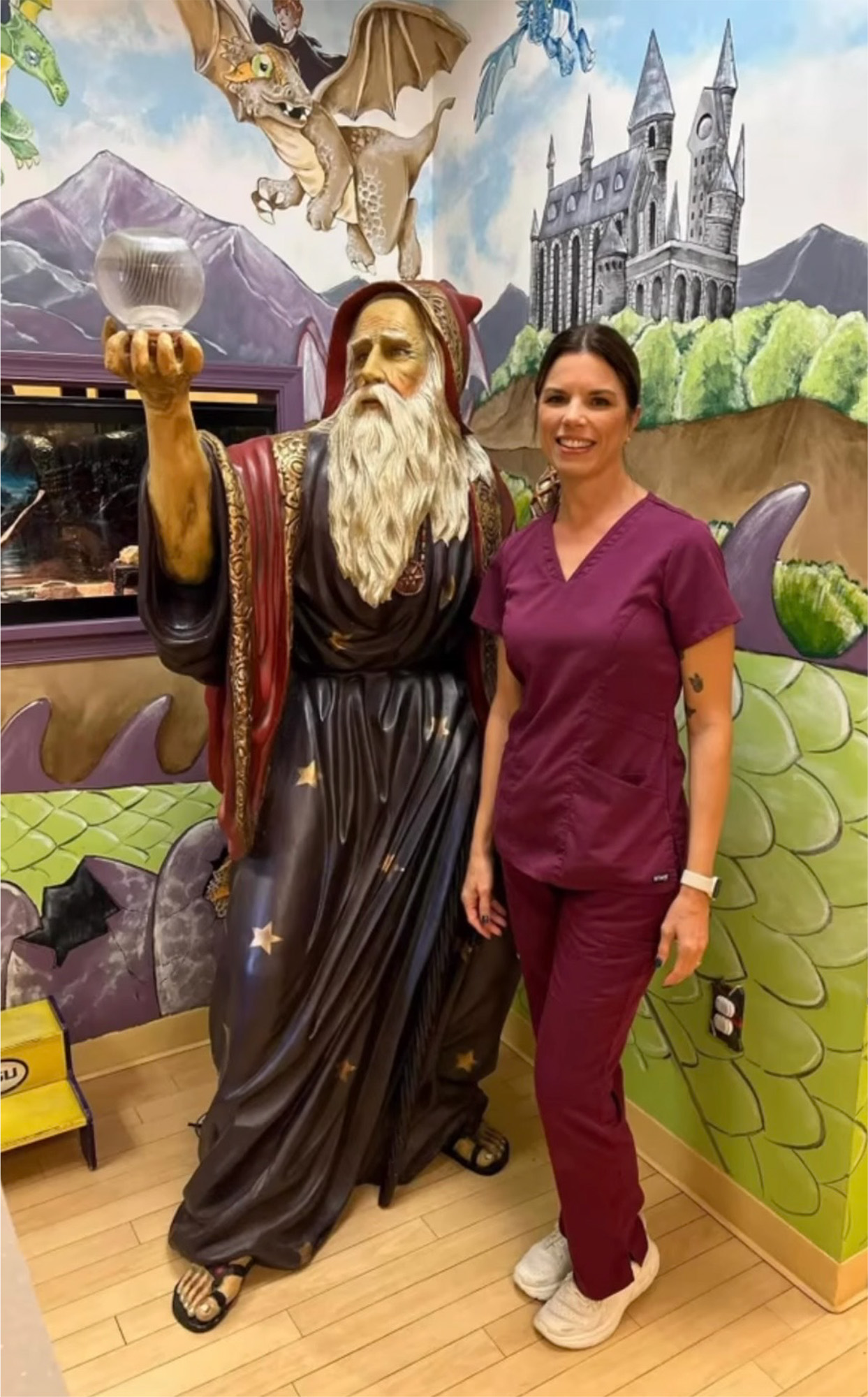 A woman in maroon scrubs stands next to a wizard statue holding a crystal ball, with colorful fantasy murals on the walls behind them—an imaginative setting for vaccination in Melbourne, FL.