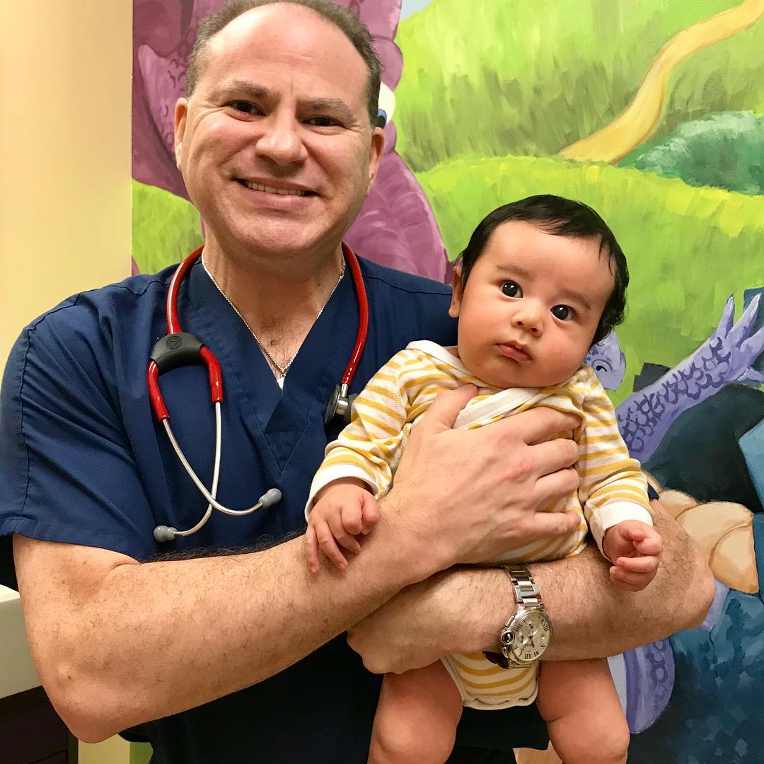 Dr. Heft, a newborn care pediatrician, in blue scrubs and a stethoscope holds an infant wearing a yellow striped outfit, highlighting attentive newborn care; a colorful mural is in the background.