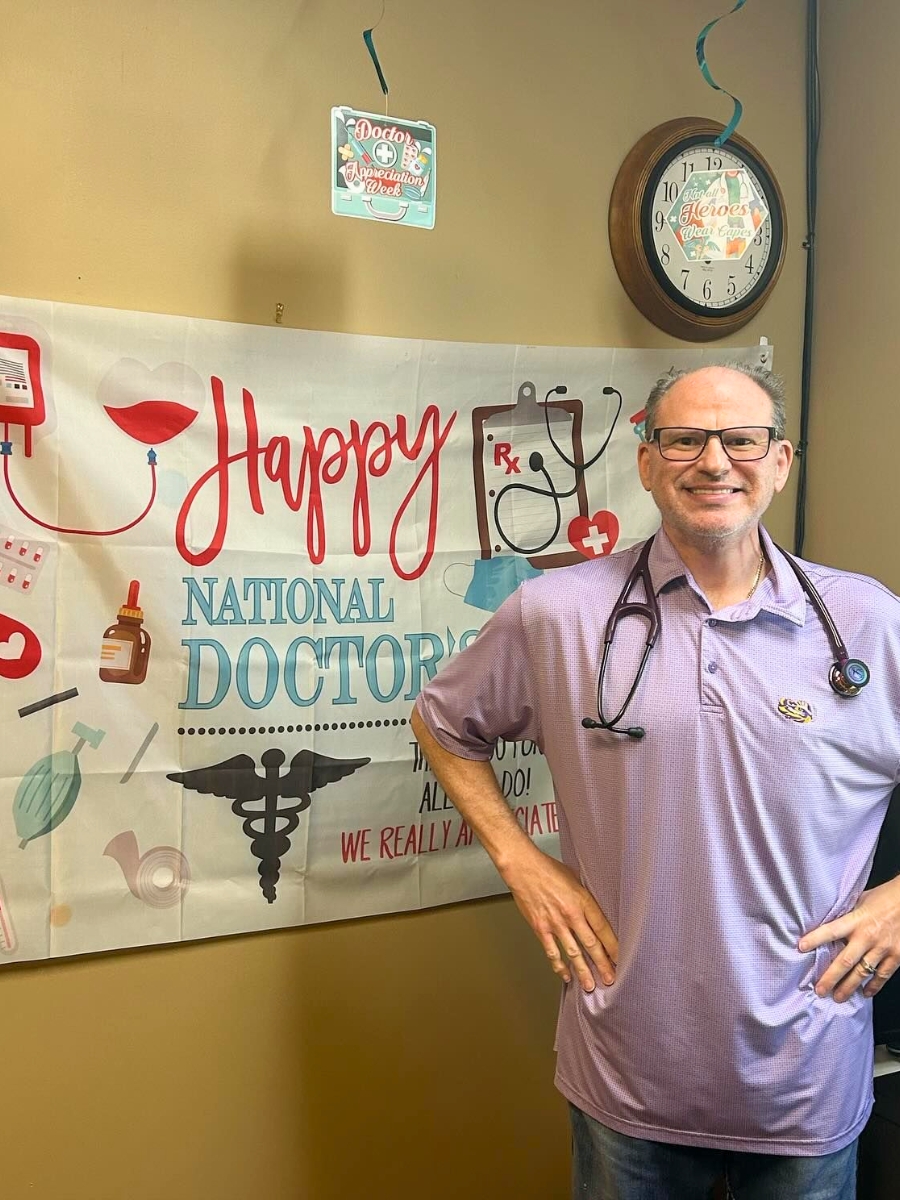 Dr. David Helft of Pediatric Wizards wearing glasses and a stethoscope stands in front of a "Happy National Doctors Day" banner and a wall clock, smiling at the camera—celebrating his dedication to Infant Care.