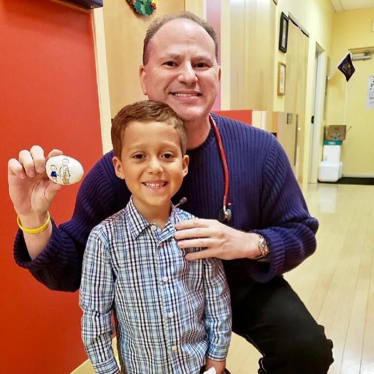 A man with a stethoscope kneels beside a smiling child holding a painted rock in a brightly lit hallway, reflecting the warmth and support of chronic care.