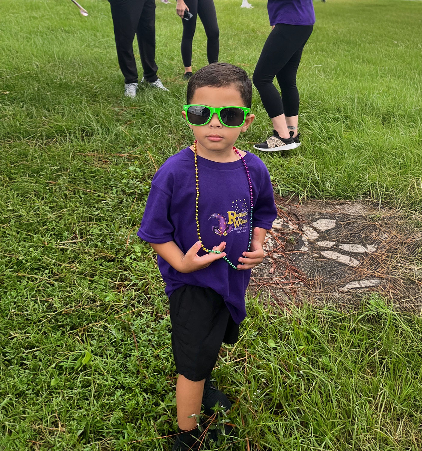 A young child in green sunglasses, a purple t-shirt, and black shorts stands on grass with a beaded necklace, while several people—possibly gathered for sports school physicals—stand behind them.