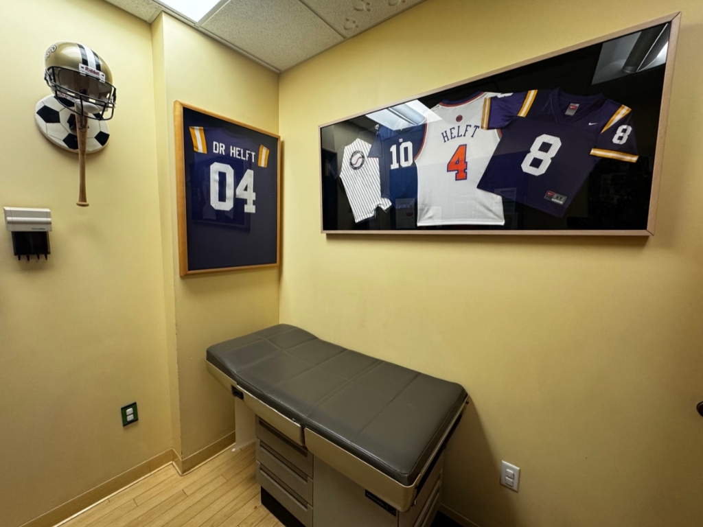 A medical exam room with a padded exam table, sports jerseys, and a football helmet mounted on beige walls reflects the welcoming atmosphere often found at a pediatrician in Melbourne, FL, all under bright overhead fluorescent lighting.
