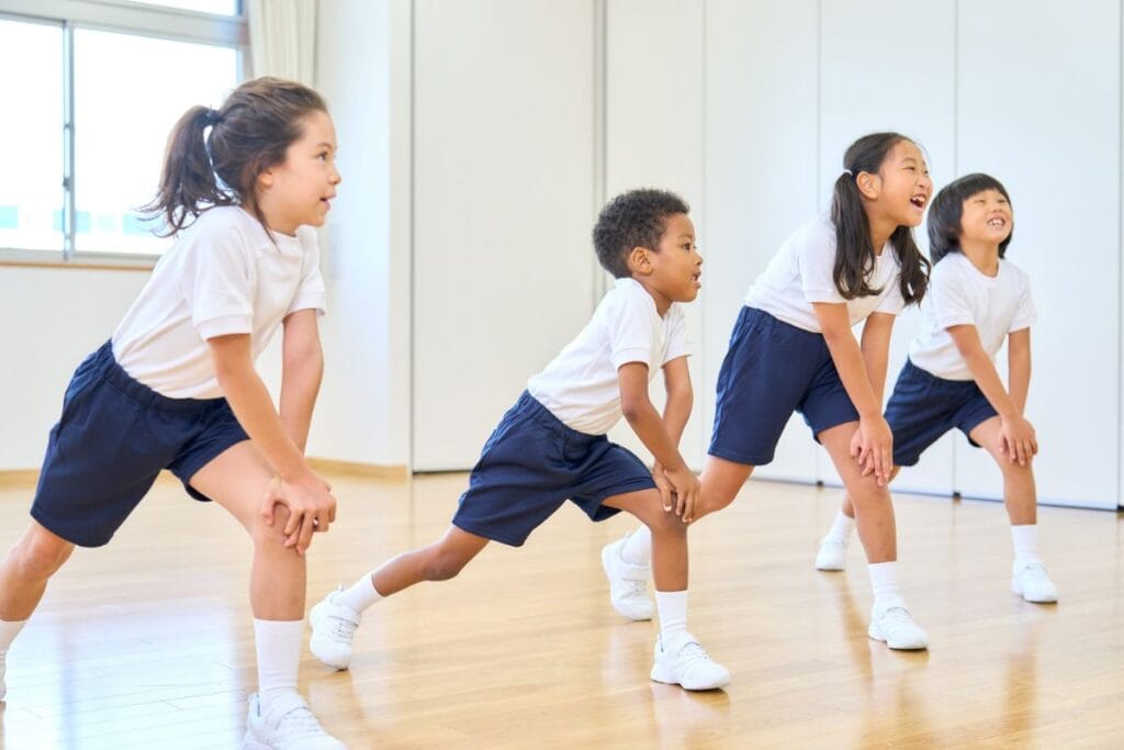 Four children in gym uniforms are standing indoors, bending forward with hands on knees, appearing to participate in a group exercise during School Sports Physicals in Melbourne FL