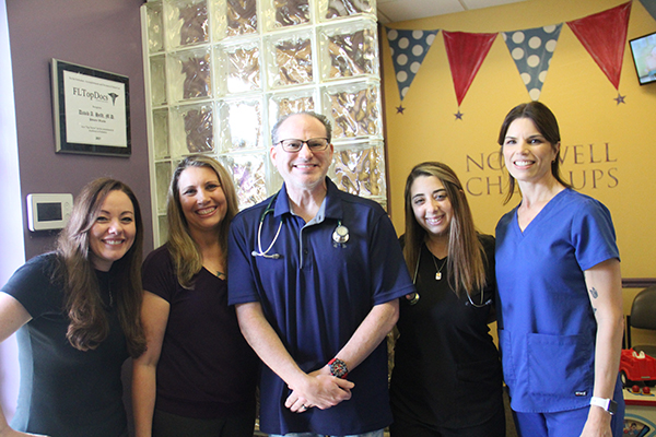 About Us: Pediatric Wizards: Five people, including three women in dark uniforms, one man in a navy shirt with a stethoscope, and one woman in blue scrubs, standing and smiling inside a clinic.