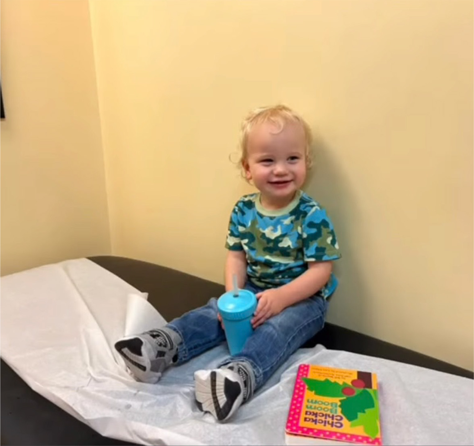 A young child in a blue camo shirt sits on an exam table holding a blue sippy cup, with a children's book beside them during their well-child checkup.