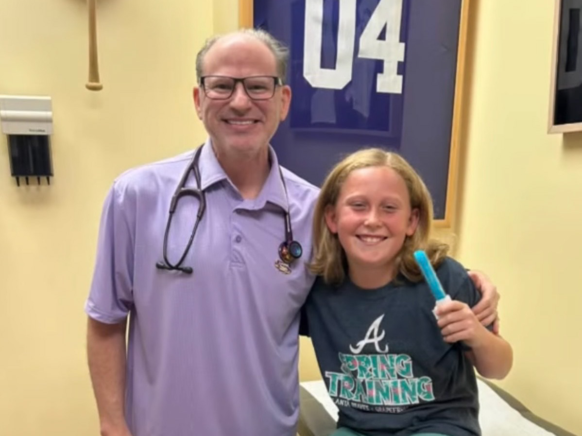 A pediatrician in Melbourne, FL with a stethoscope stands next to a smiling child holding a tongue depressor in a medical office.