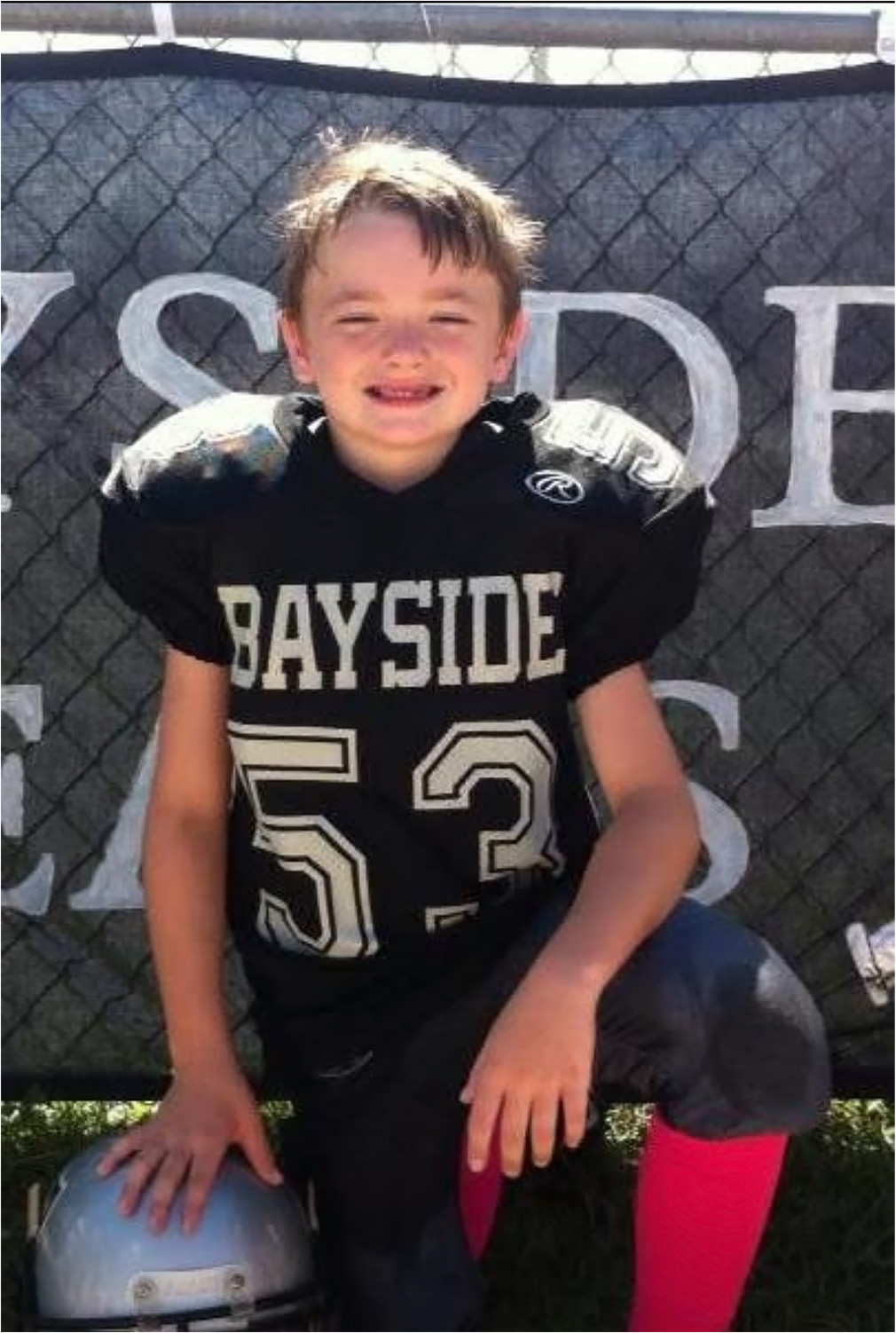 A young football player in a black Bayside jersey with the number 53 kneels on one knee, posing with a helmet in front of a fence, ready for action after passing his sports school physicals.