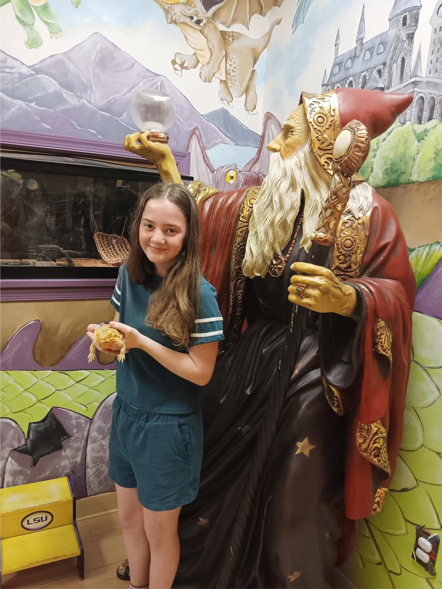 A young girl stands next to a large wizard statue holding a crystal ball, with a fantasy-themed mural in the background—turning her well-child checkups into a magical adventure.
