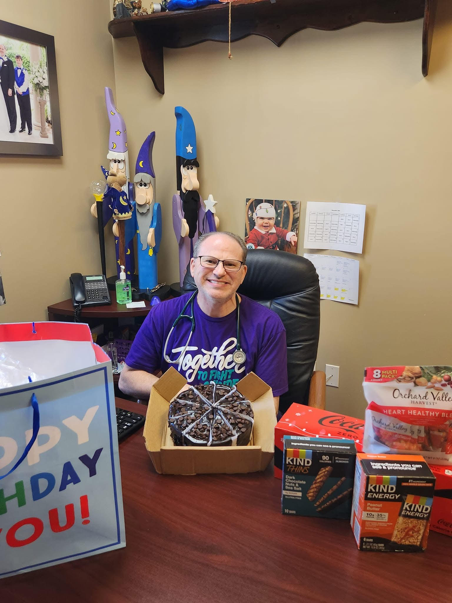 A man sits at a desk with a birthday cake, snacks, and gifts, smiling in his "Together to Flourish" shirt. Wizard figurines and photos decorate the office, creating a festive vibe perfect for celebrating after getting a vaccination in Melbourne FL.