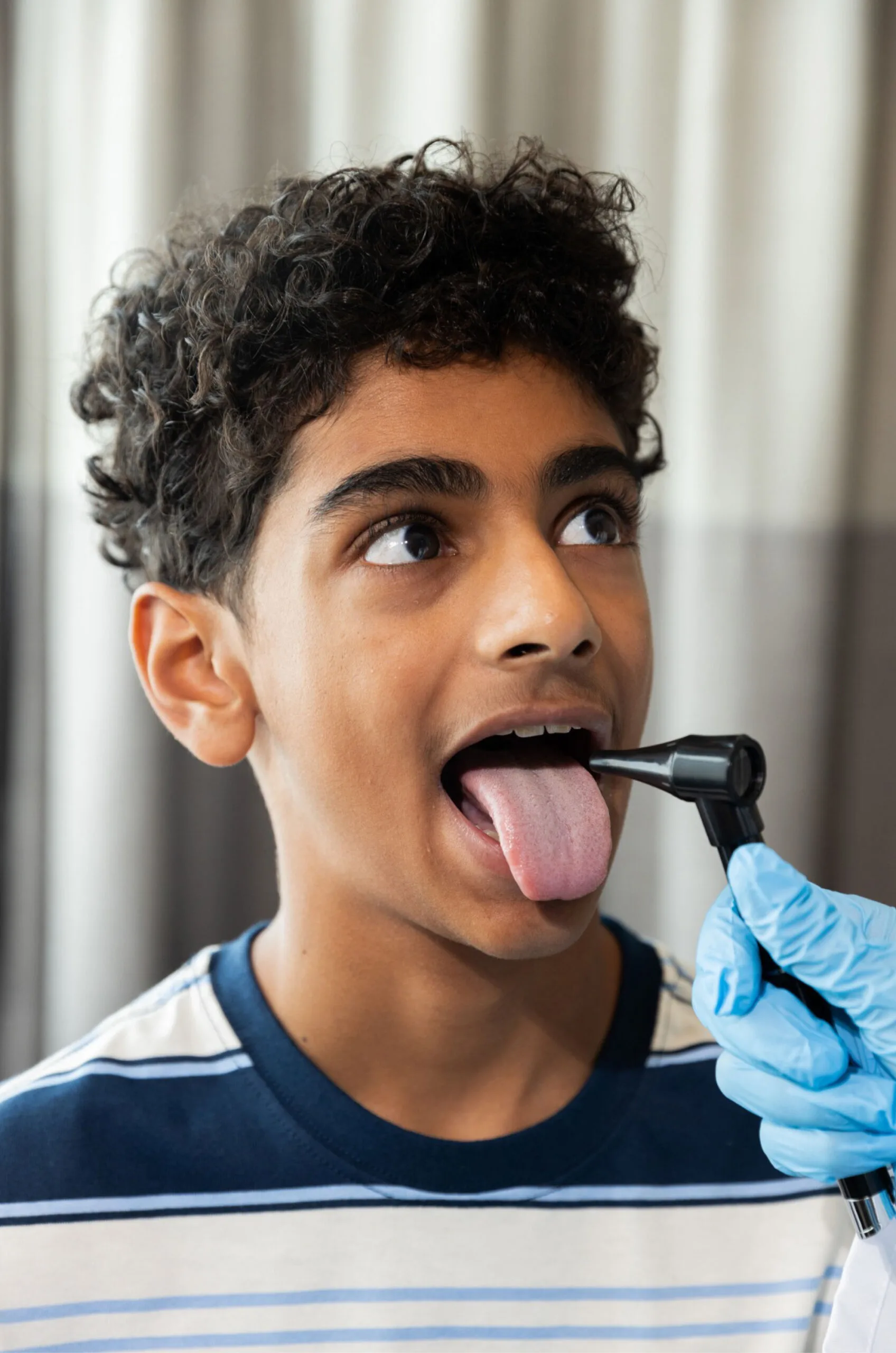 A healthcare professional in blue gloves examines a boy&rsquo;s throat with a medical instrument, while nearby, his insurance information is reviewed.