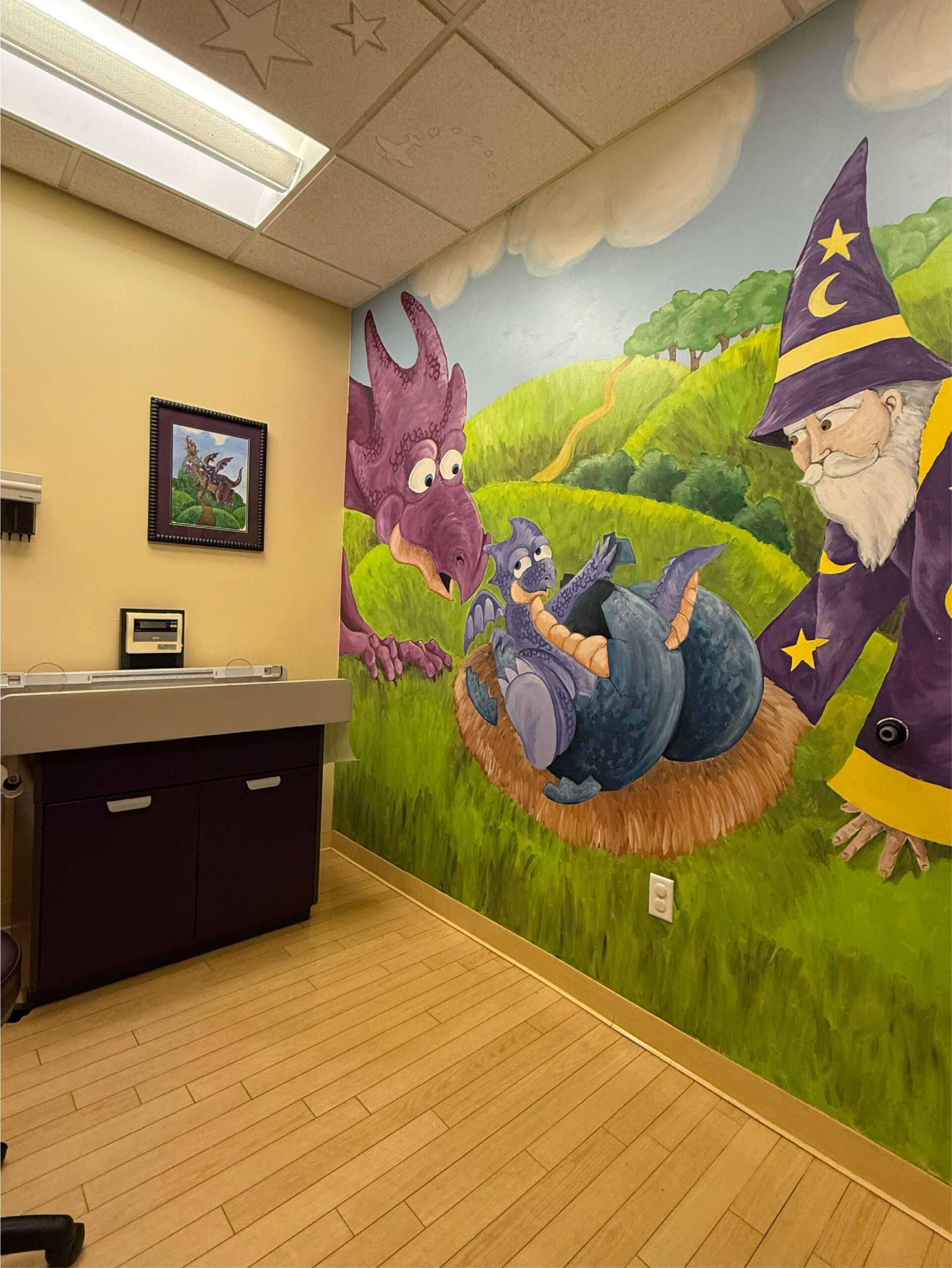 A pediatric exam room designed for newborn care features a wall mural with a purple dragon, blue dragon, and wizard in a hilly landscape; framed art hangs above the left counter.