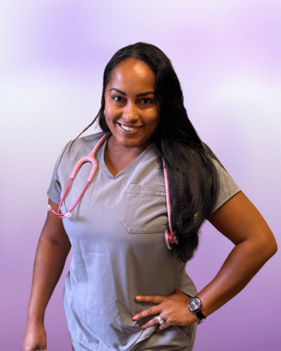 Woman wearing gray scrubs and a pink stethoscope smiles at the camera, standing with one hand on her hip against a light purple background—meet the face behind our about us story.